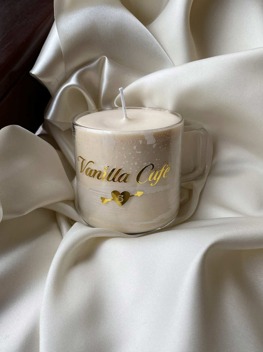 Vanilla Café is a scent candle that has notes of dreamy vanilla and espresso. The candle is designed to look like a cup of offee, laying on a bed of beige satin fabric.