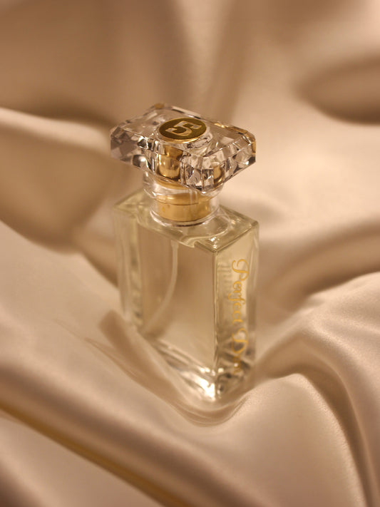 a clear perfume bottle with a crystal like cap with a golden Logo that reads "Perfect Dim" standing on a golden satin fabric.