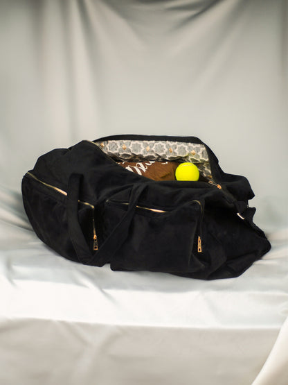 A black matte velvet tennis bag with golden flower details, open, displayed against a cream white satin background.