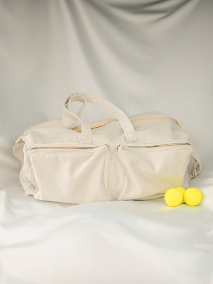 a closed view of the cream white velvet tennis bag with golden zippers and two front pockets. Standing on a golden satin background.